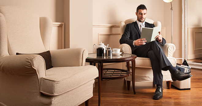 A man sitting on one of the two chairs reading a book and a table is placed in between the two chairs
